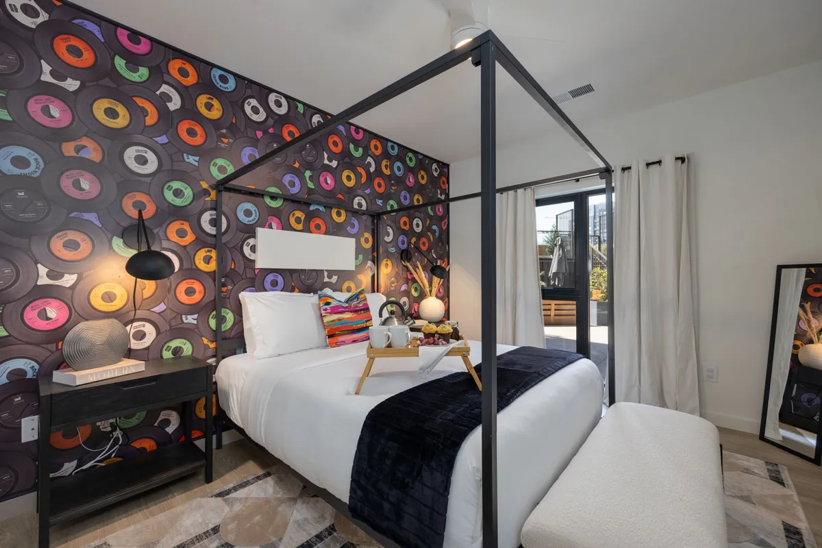 A vibrant bedroom featuring a canopy bed adorned with colorful record wallpaper as a backdrop, a stylish bedside lamp, and a breakfast tray resting on the neatly made bed. The decor exudes musical energy and creativity, while the setting feels inviting for relaxation and morning indulgence.