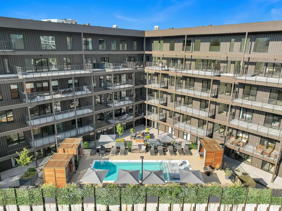 An exterior view of a lively communal swimming pool surrounded by seating areas, balconies, and lush landscaping. The atmosphere is vibrant yet relaxed, suggesting group gatherings, socializing, and outdoor fun.