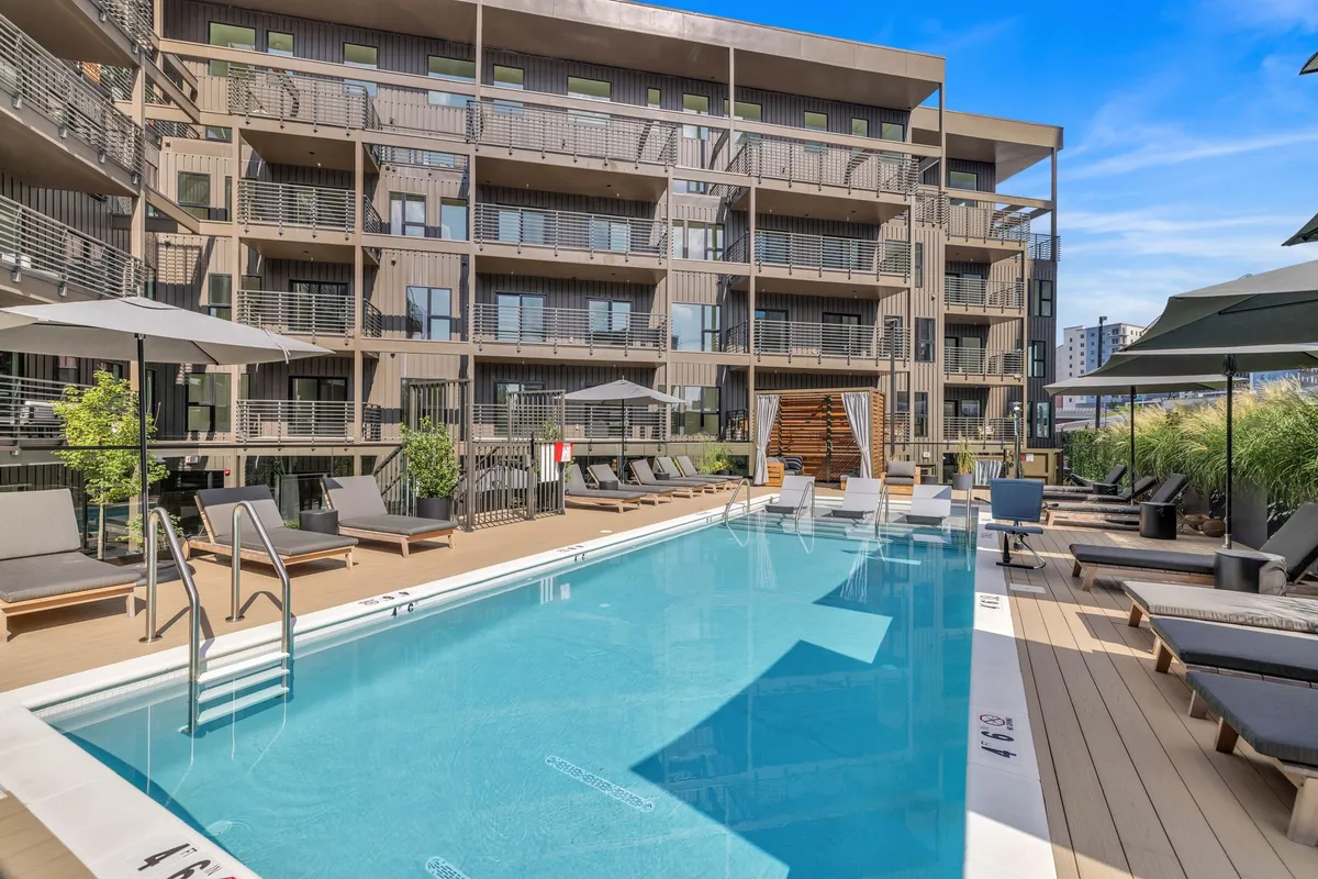 A sunlit swimming pool lined with comfortable lounge chairs and surrounded by lush greenery. The sleek building facade can be seen in the background, suggesting a modern and inviting environment perfect for group relaxation or socializing.