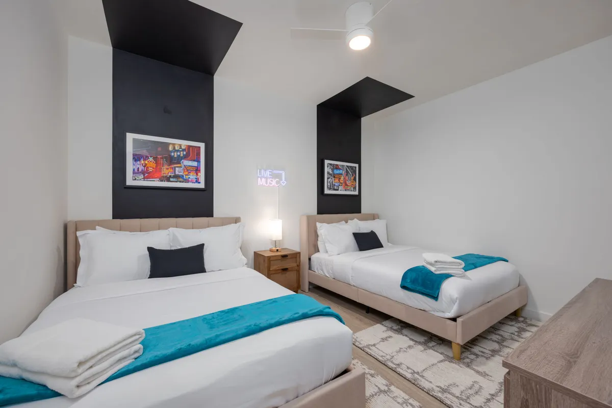 A second bedroom with two beds accented by turquoise touches, framed art on the walls, and a prominent neon sign reading 'LIVE MUSIC.' A wooden nightstand with a lamp sits between the beds, creating an inviting, energetic space for guests to unwind or share stories.