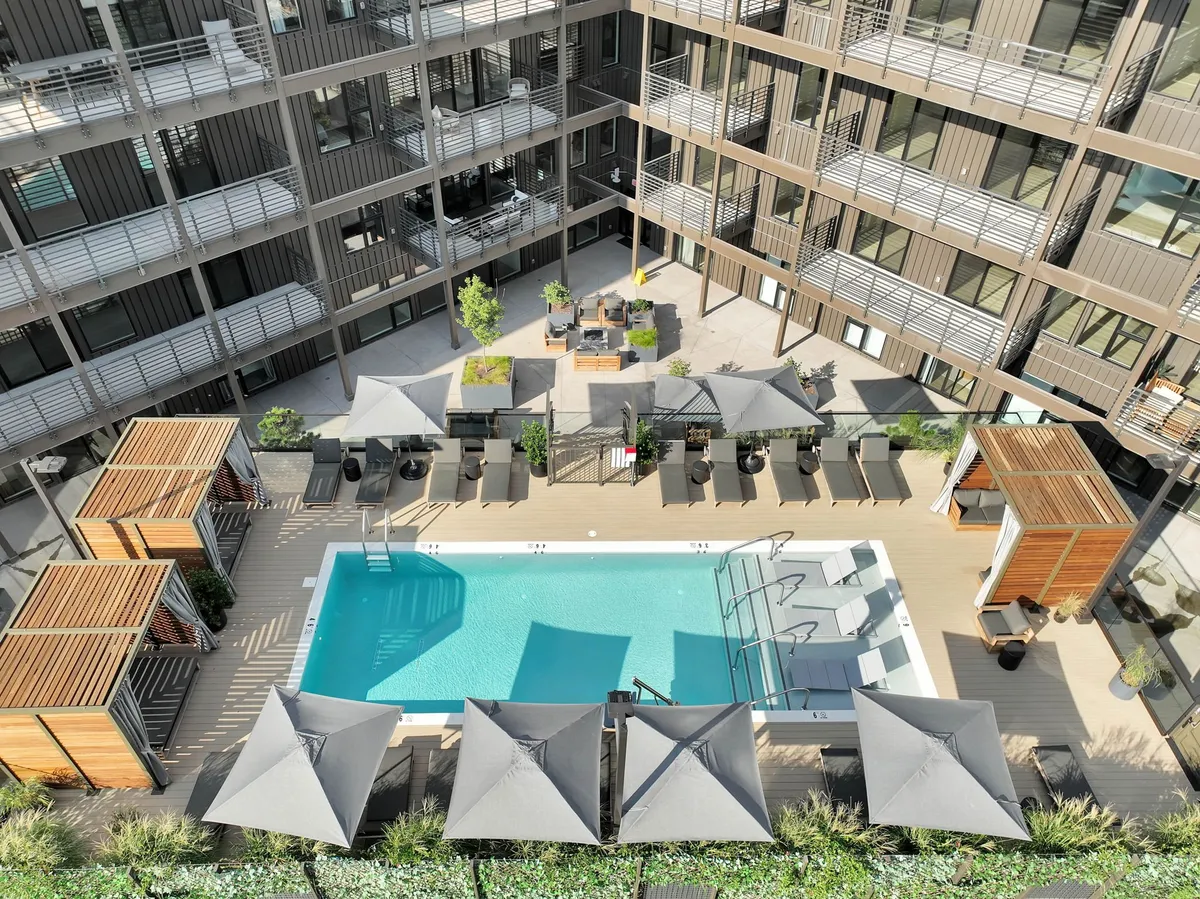 This image depicts a sparkling outdoor swimming pool surrounded by lounge chairs, umbrellas, and landscaped areas. The setting is tranquil, with hints of sunset lighting and a deck area that could host barbecues or morning yoga. Groups of friends are seen enjoying the space, suggesting a vibrant community atmosphere.