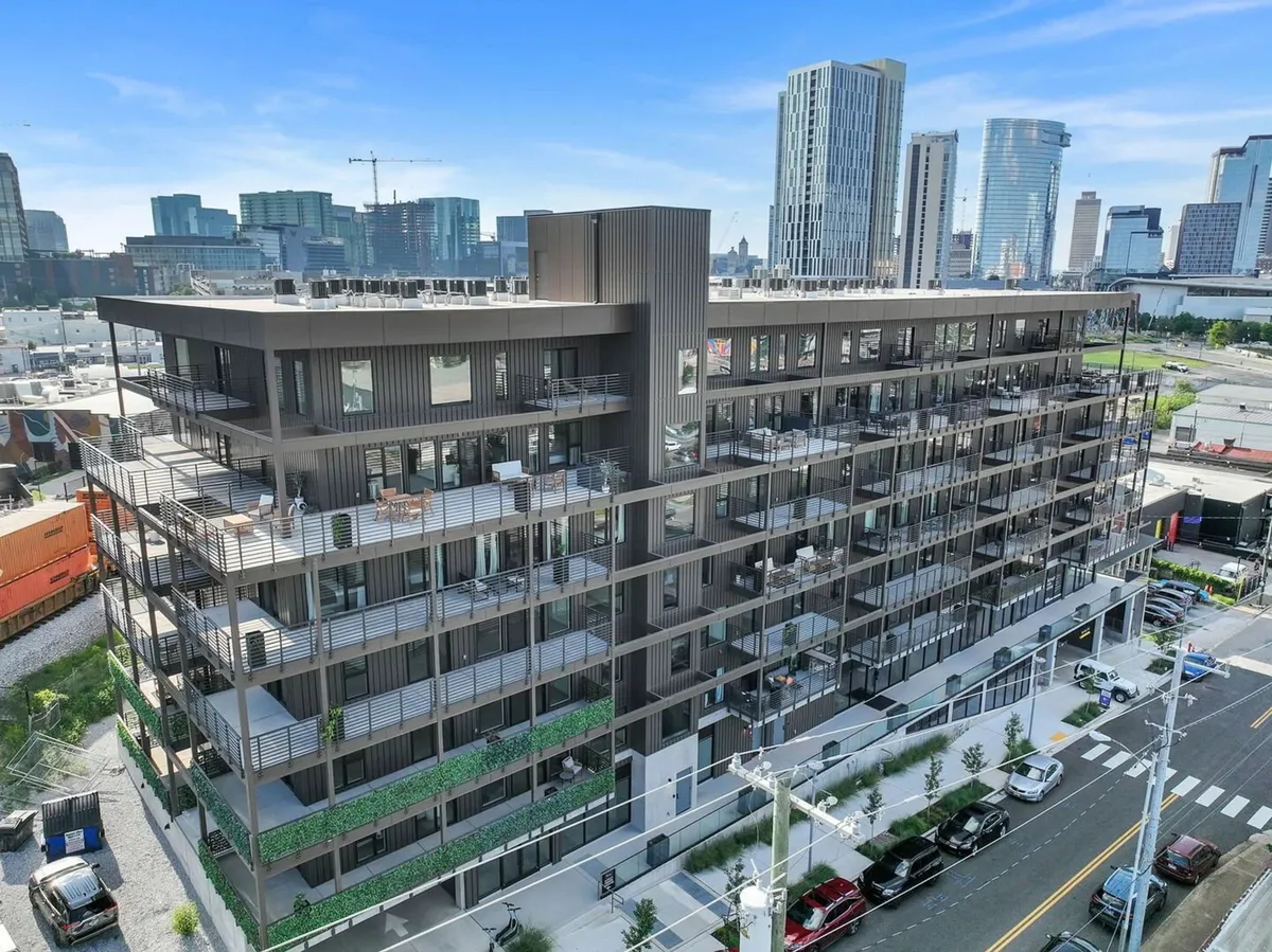 This image showcases a modern apartment building with a rooftop terrace overlooking the city skyline, set against a vibrant street view with parked cars. The architecture suggests an energetic, urban environment right in the heart of downtown Nashville.