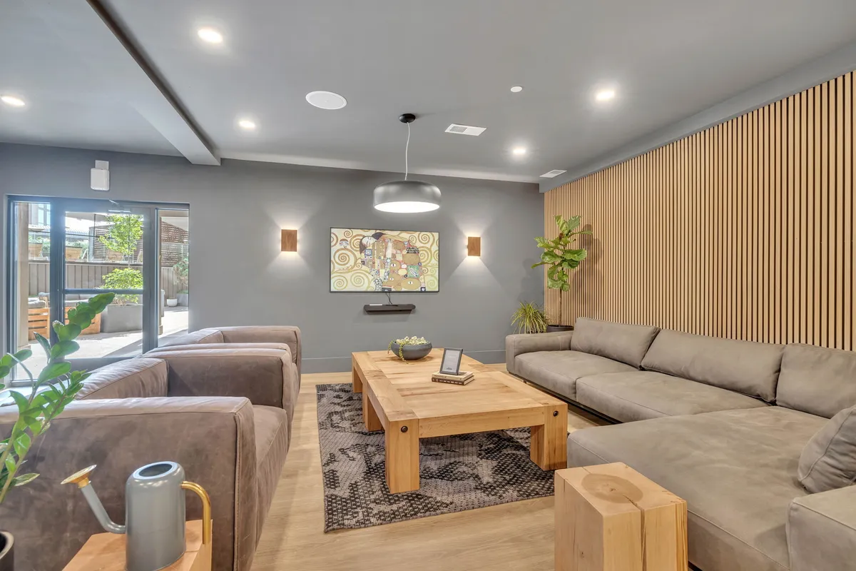 A spacious, open living area featuring a large sectional sofa, a wooden coffee table, vibrant artwork on the wall, and potted plants in the corners. The room is arranged to facilitate group gatherings and casual conversations, with direct sightlines hinting at an open-concept layout.