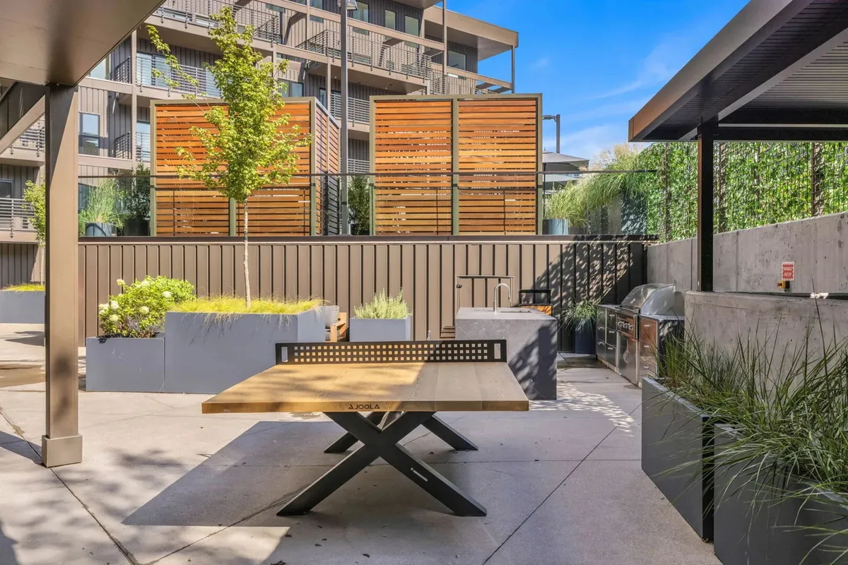 The image showcases an outdoor barbecue area with a wooden table, privacy screens, surrounding greenery, and a relaxed, welcoming ambiance. People are gathered for outdoor dining, capturing the spirit of communal meals and socializing with friends.
