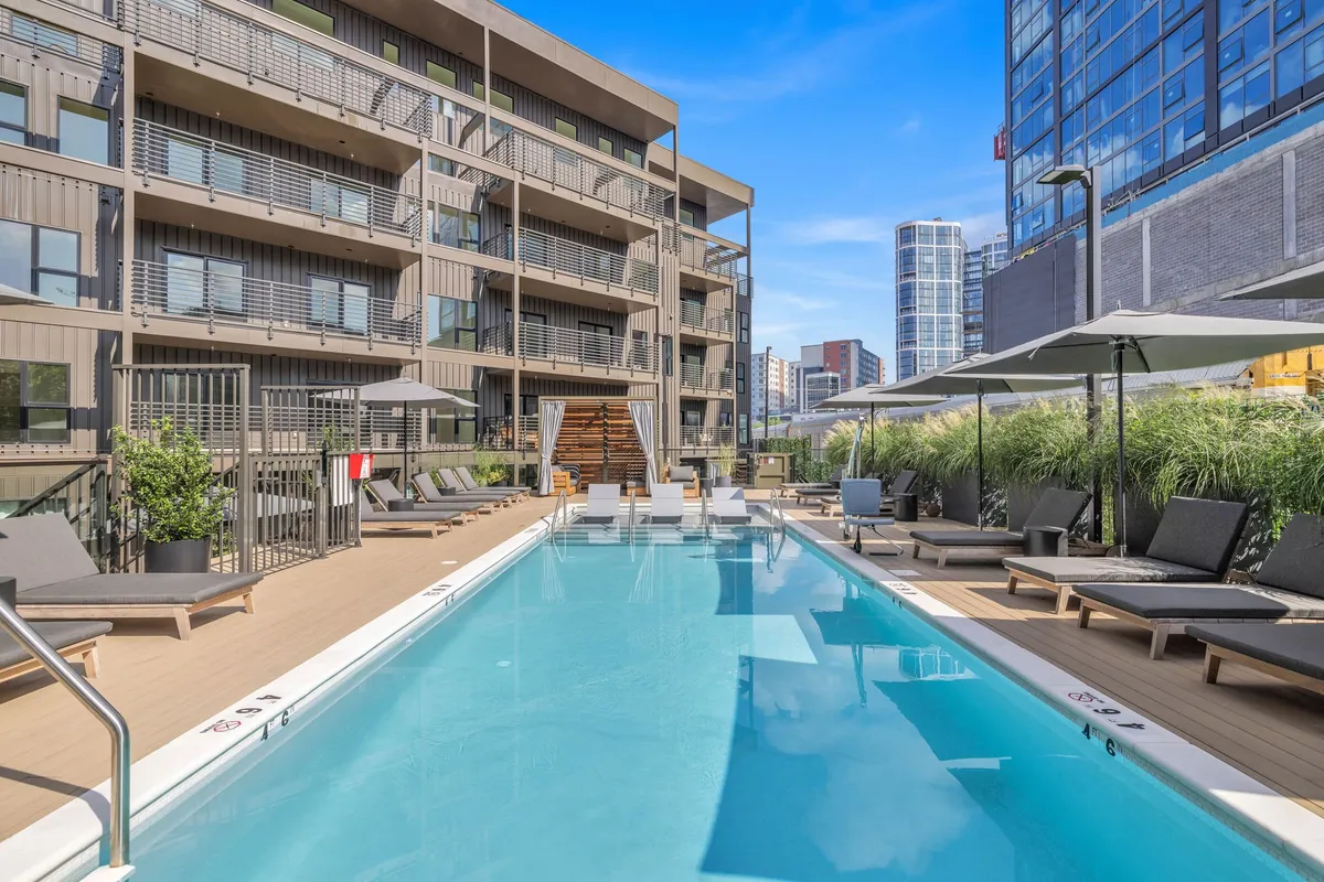 A vibrant exterior pool area featuring lush landscaping, inviting lounge chairs, and a sparkling swimming pool surrounded by greenery. The space exudes a relaxed social ambiance, suggesting activities like sunbathing, swimming, and gathering with friends. The building facade and the warm glow of sunset add to the resort-inspired vibe.